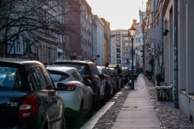Une rue en ville. Un certain nombre de voitures se garent à côté d'un trottoir étroit. Il y a peu de gens à voir.