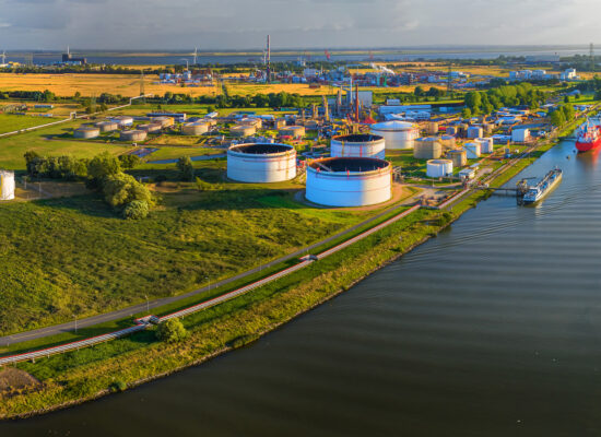 Drohnenansicht eines Industriekomplex mit Gas, Öl und Wasserstoffspeichern, Windräder im Hintergrund.