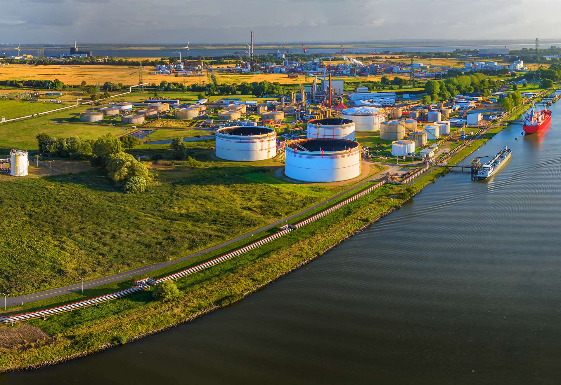 Drohnenansicht eines Industriekomplex mit Gas, Öl und Wasserstoffspeichern, Windräder im Hintergrund.